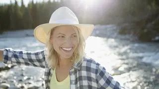 Kobieta w koszuli w kratę i kapeluszu na świeżym powietrzu. W tle jezioro.