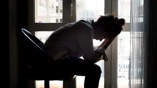 Załamana kobieta siedzi na krześle na tle okna. Trzyma głowę w dłoniach.