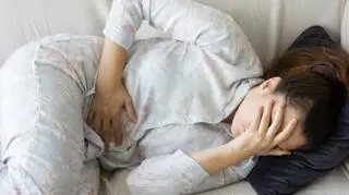 mloda kobieta lezy na kanapie trzyma sie za brzuch trzyma reke przy twarzy zmartwiona