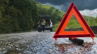 Trójkąt ostrzegawczy na pierwszym planie. W tle zepsuty samochód. 