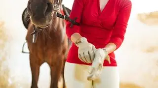 Kobieta zakłada rękawice jeździeckie, w tle stoi koń