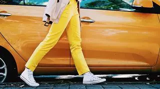Kobieta w żółtych materiałowych spodniach i trampkach na tle samochodu. 