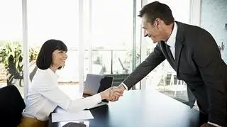 Mężczyzna ściskka siedzącej za biurkiem kobiecie dłoń. 