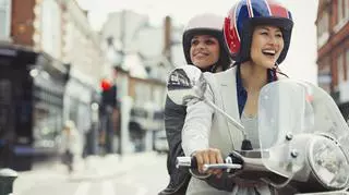 Szczęśliwi ludzie jadą skuterem lub motorowerem przez ulice miasta. Mają kaski na głowach.