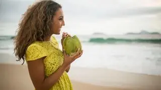 Kobieta w żółtej sukience nad brzegiem morza, pijąca wodę z kokosa