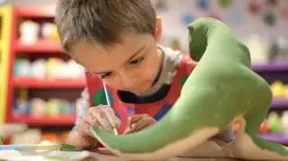 maly chlopiec siedzi przy stole przed figurka dinozaura rysuje