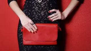 Kobieta w czarnej koronkowej sukience, z czerwoną torebką w dłoni. 