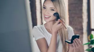 Jasnowłosa kobieta poprawiająca makijaż przed lustrem.