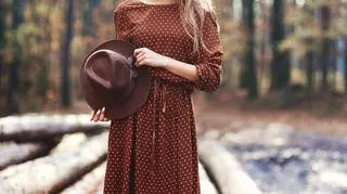 Kobieta w brązowej sukience trzyma w ręku kapelusz