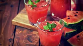 Dwie szklanki z czerwonym sokiem ozdobione listkami mięty, obok pokrojony owoc arbuza.