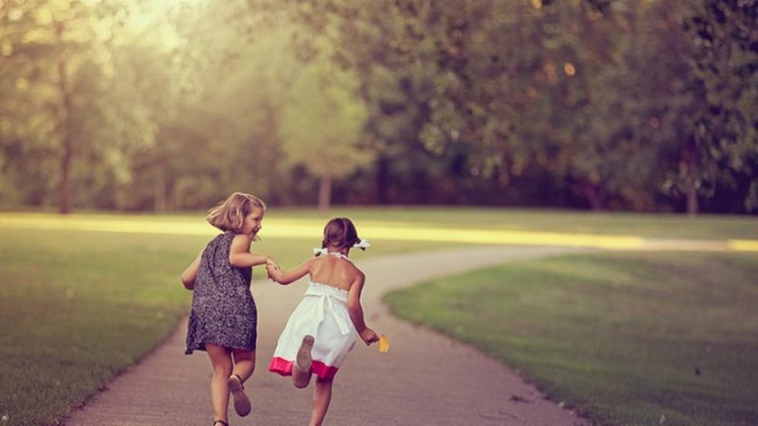 2 dziewczynki w sukienkach biegną przez park i trzymają się za ręce