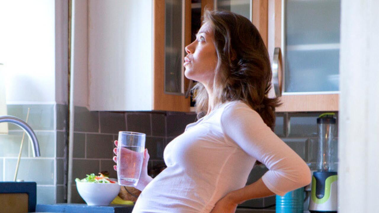 kobieta w zaawansowanej ciazy stoi w kuchni ze szklanka oddycha gleboko trzyma sie za biodra