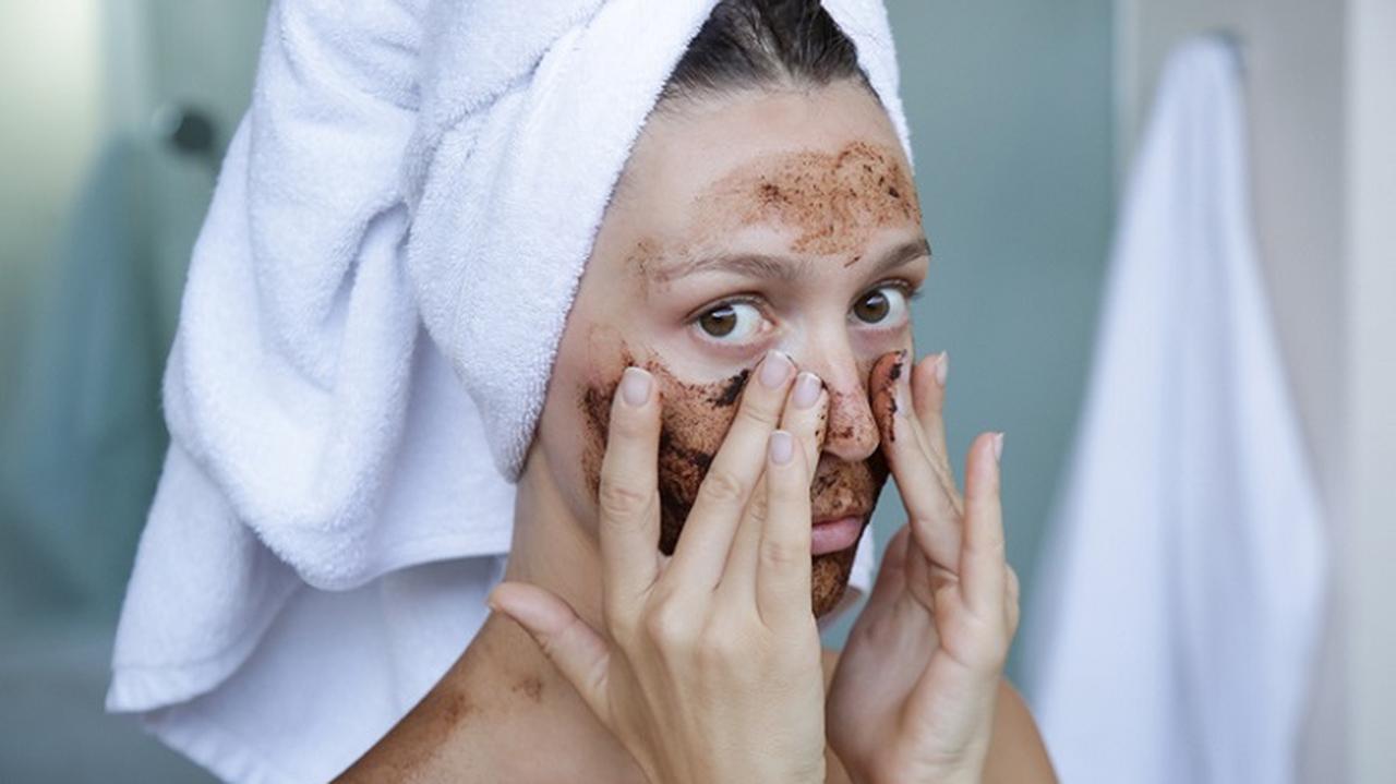 kobieta w białym ręczniku na głowie nakłada peeling kawowy na twarz