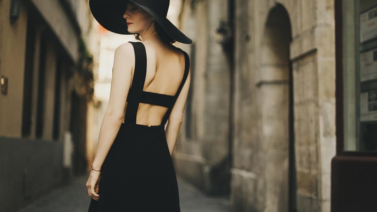 Kobieta w czarnej seksownej sukience i kapeluszu idzie ulicą. 