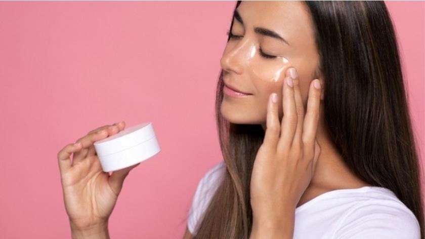 Kobieta nakładająca krem na twarz, w dłoni trzymająca słoiczek z kosmetykiem