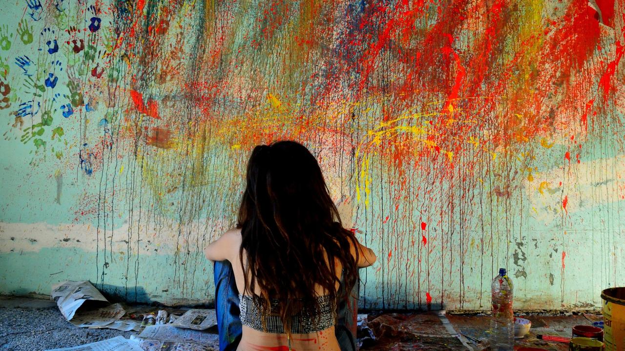 Dziewczyna siedzi i patrzy na ścianę zachlapaną kolorowymi farbami ścianę