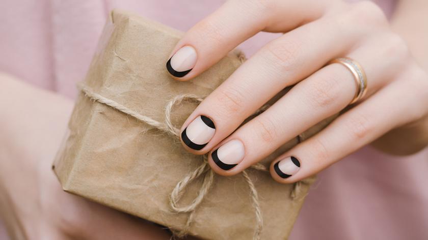 Kobieca dłoń trzymająca owinięte szarym papierem pudełko. Na paznokciach czarny french manicure - modna technika zdobienia