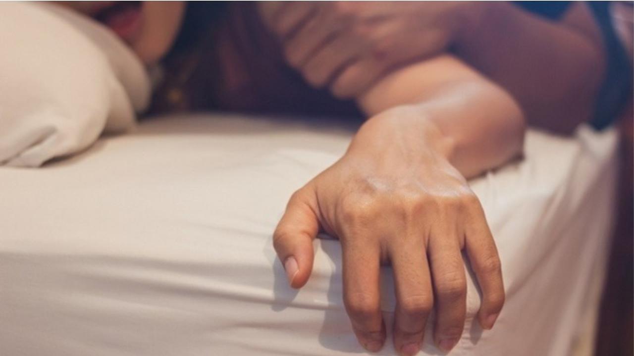 Kobieca dłoń kurczowo trzymająca materac łóżka, w tle twarz jej właścicielki i jej ciało, przytrzymywane męskim ramieniem