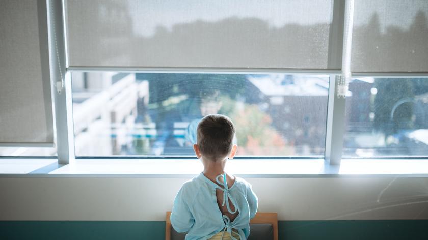 Chłopiec w szpitalnym ubraniu patrzy przez okno. 