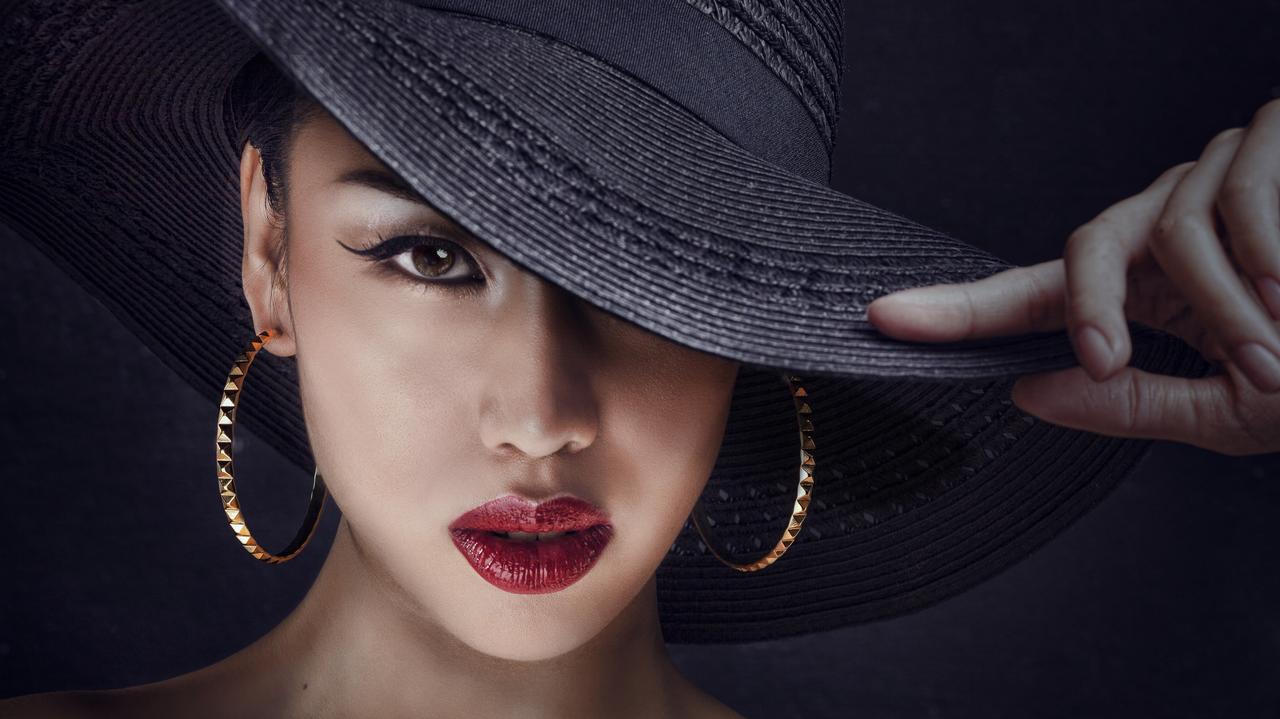 Elegancka kobieta z wyrazistym makijażem. Na jej głowie duży czarny kapelusz.