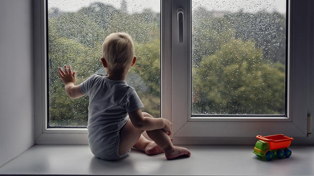 Dziecko siedzi na parapecie i patrzy przez okno w deszczową pogodę
