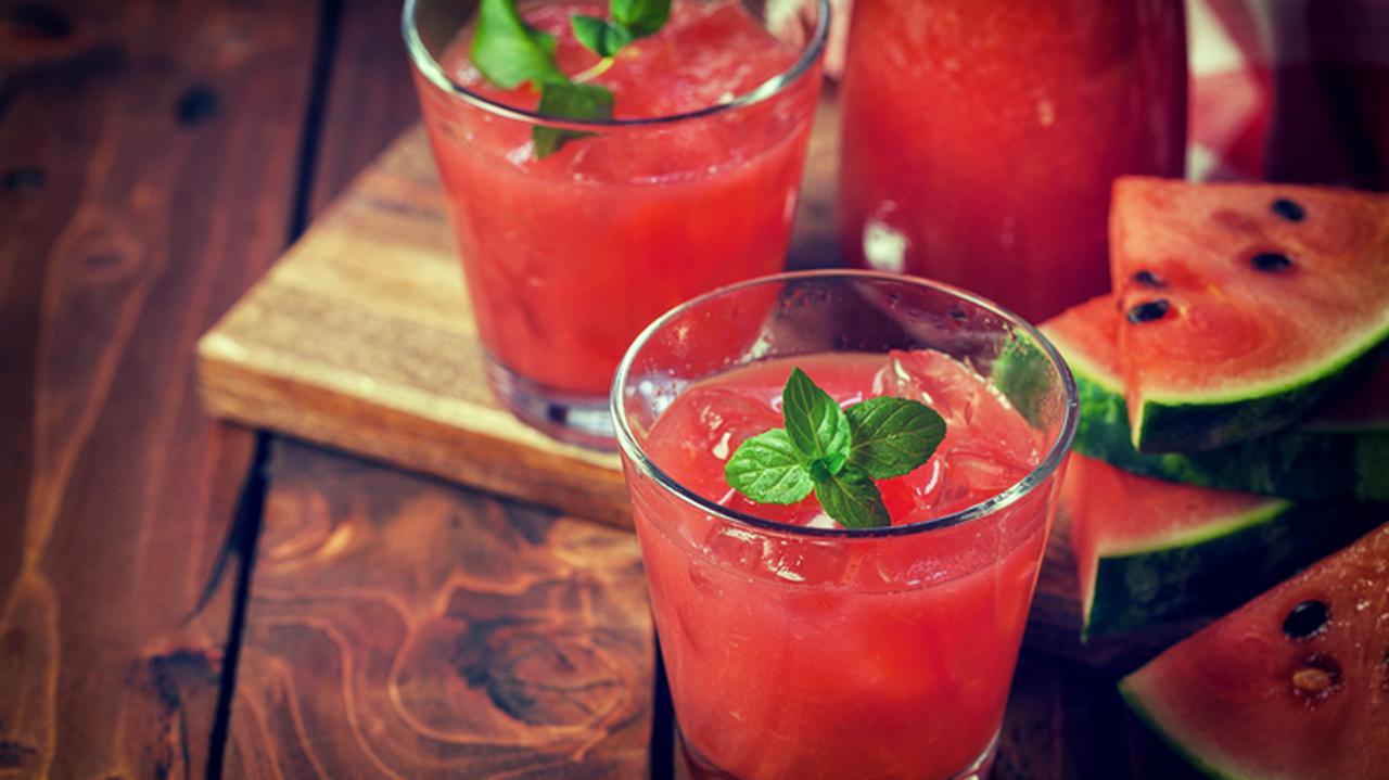 Dwie szklanki z czerwonym sokiem ozdobione listkami mięty, obok pokrojony owoc arbuza.